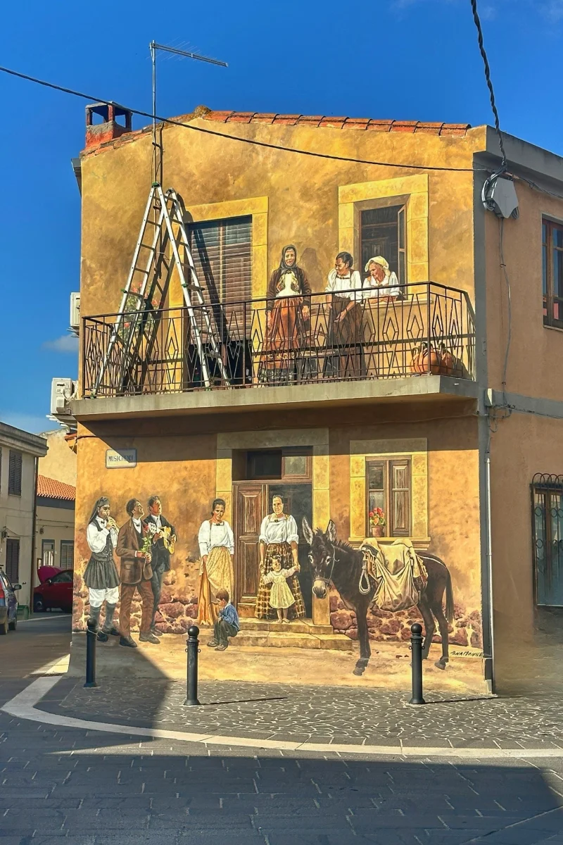 Murales auf Sardinien: Wandgemälde auf ockerfarbener Hauswand mit Dorfbewohnern in traditionellen Trachten und Esel.