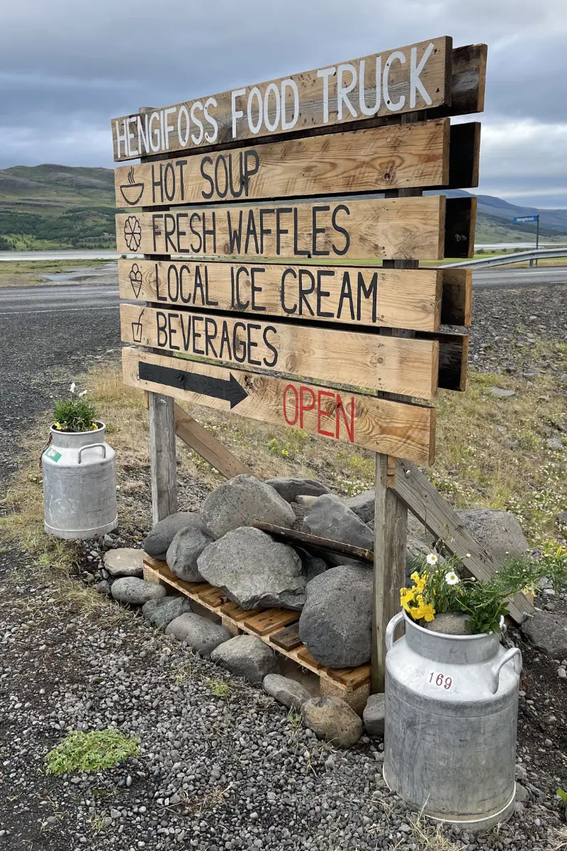 Rustikales Holzschild des Hengifoss Food Trucks in Island, das Waffeln, Eis und Suppe anbietet; flankiert von Milchkannen.