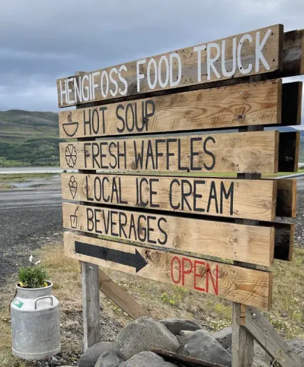 Rustikales Holzschild des Hengifoss Food Trucks in Island, das Waffeln, Eis und Suppe anbietet; flankiert von Milchkannen. Rustikales Holzschild des Hengifoss Food Trucks in Island, das Waffeln, Eis und Suppe anbietet; flankiert von Milchkannen.