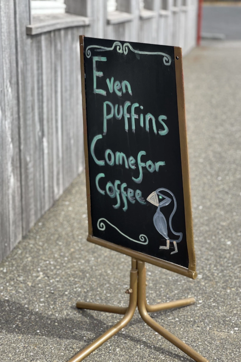Kreidetafel-Aufsteller vor Holzfassade mit der Aufschrift Even puffins Come for Coffee und kleiner Papageientaucherzeichnung.
