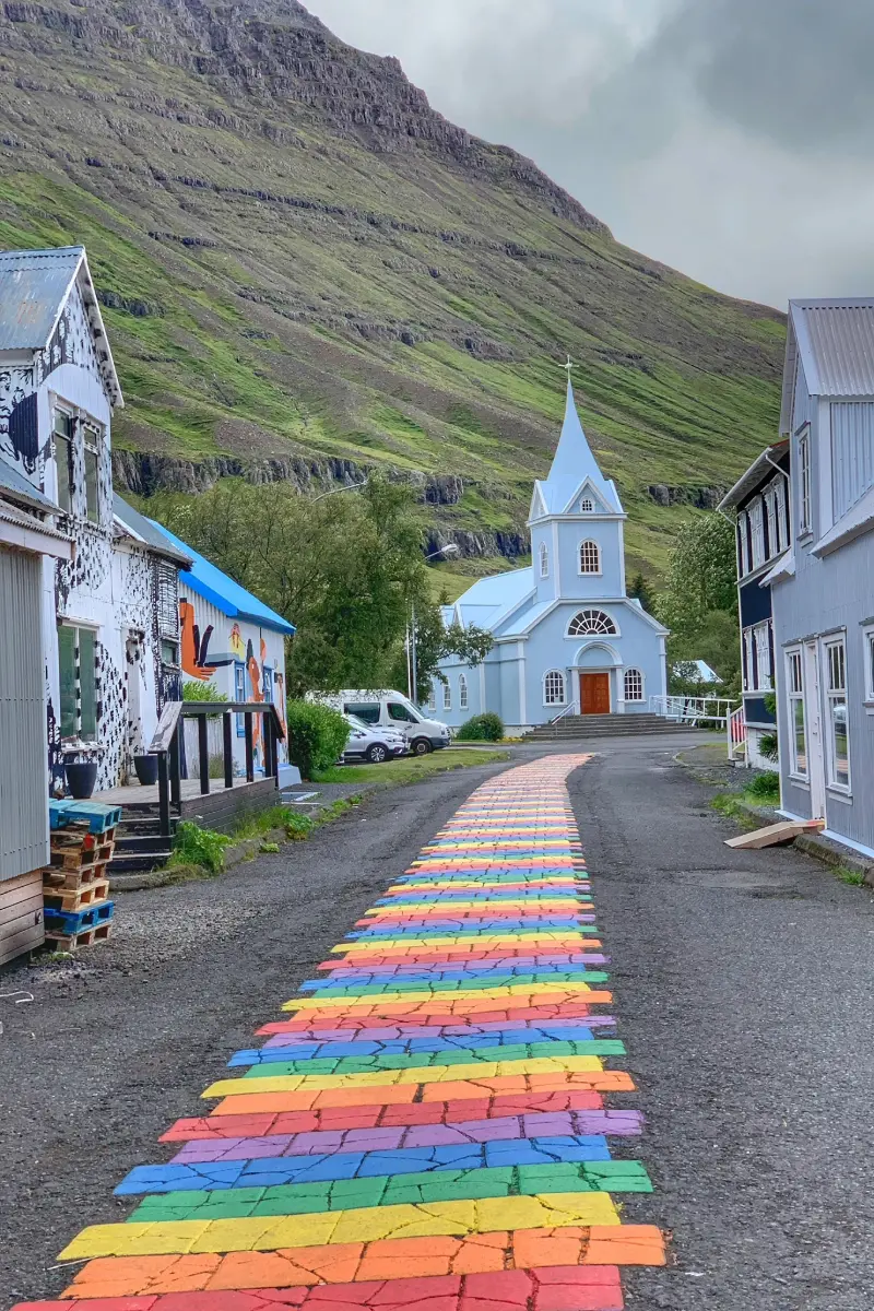 Regenbogenweg führt zur berühmten hellblauen Seyðisfjarðarkirkja-Kirche in Seyðisfjörður vor steilen Berghängen Islands.