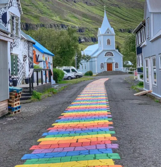 Regenbogenweg führt zur berühmten hellblauen Seyðisfjarðarkirkja-Kirche in Seyðisfjörður vor steilen Berghängen Islands. Regenbogenweg führt zur berühmten hellblauen Seyðisfjarðarkirkja-Kirche in Seyðisfjörður vor steilen Berghängen Islands.