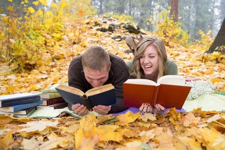 Owners, man and woman reading books and smiling while sitting on a bed of golden leaves