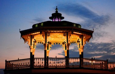 Side view of yellow lit Brighton Bandstand in the night in blue background sky. Photo by Eva Kalpadaki. Side view of yellow lit Brighton Bandstand in the night in blue background sky. Photo by Eva Kalpadaki.