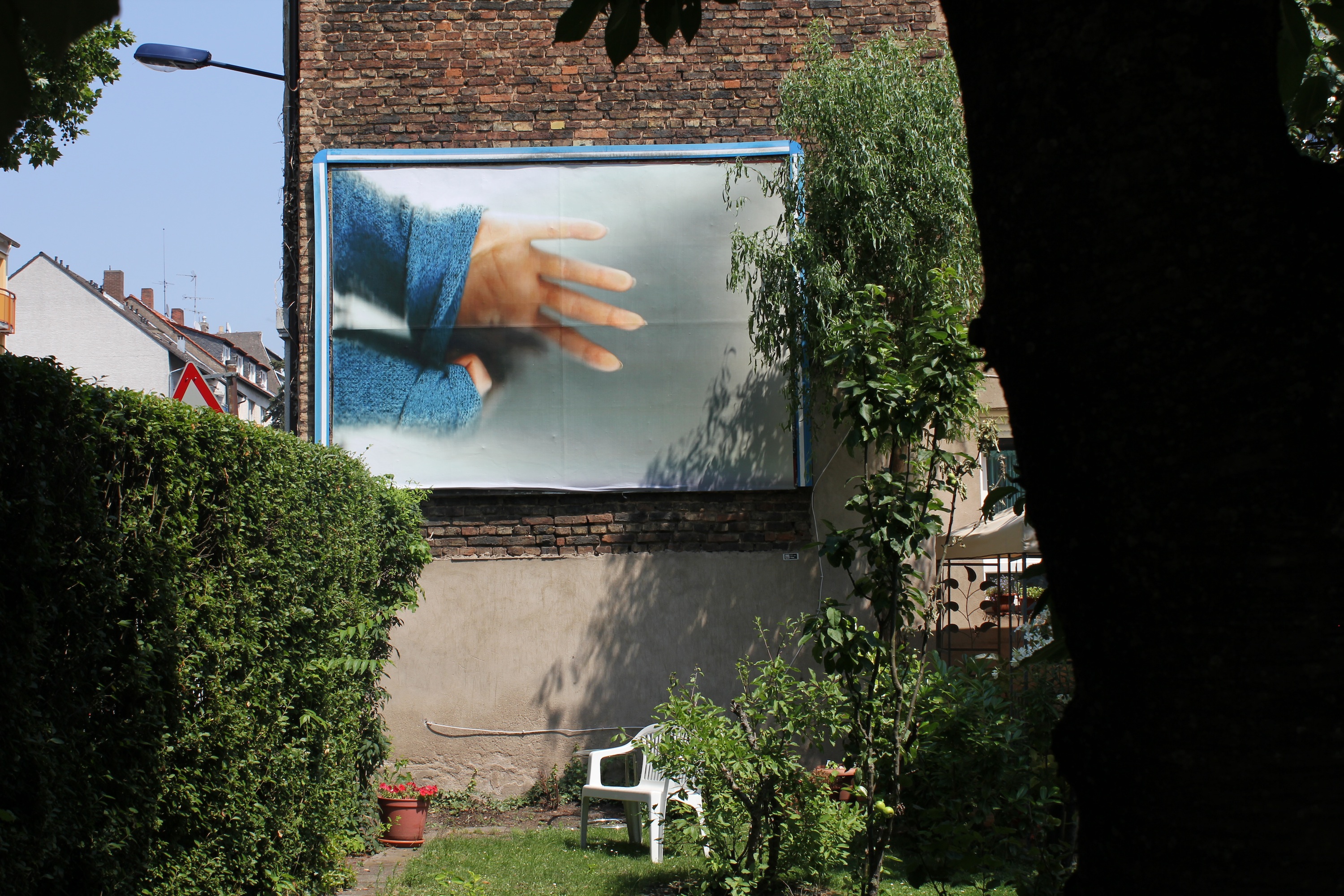 billboard inside a garden. The billboard image is of an arm with a blue jumper and a hand pulling at it. The background is a brick wall and a hedge. The garden is green, with a white deck chair, potted plants and trees