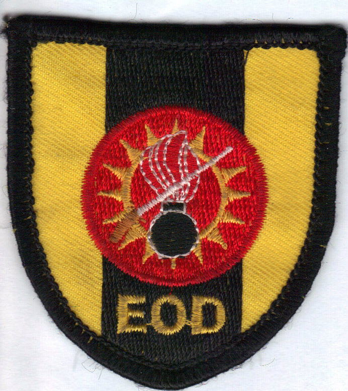 AMMUNITIONSRYDNINGSCENTERENE
Autoriseret:
4. januar 1985	 
Anlagt:
1. maj 1988	
Motiv:
En rød cirkel hvori strålekrans med brændende bombe og sabel, alt skjoldlagt.	
Symbolik:
Den røde cirkel symboliserer ild og eksplosion. Den gule strålekrans refererer til personellets evne til at fortage bortsprængning af alle former for ammunition. Den brændende bombe, hvis flamme er gennemskåret af en ingeniørsabel M/1866, refererer til personellets kunnen med hensyn til at afbryde tændforløbet i en ammunitionsgenstand