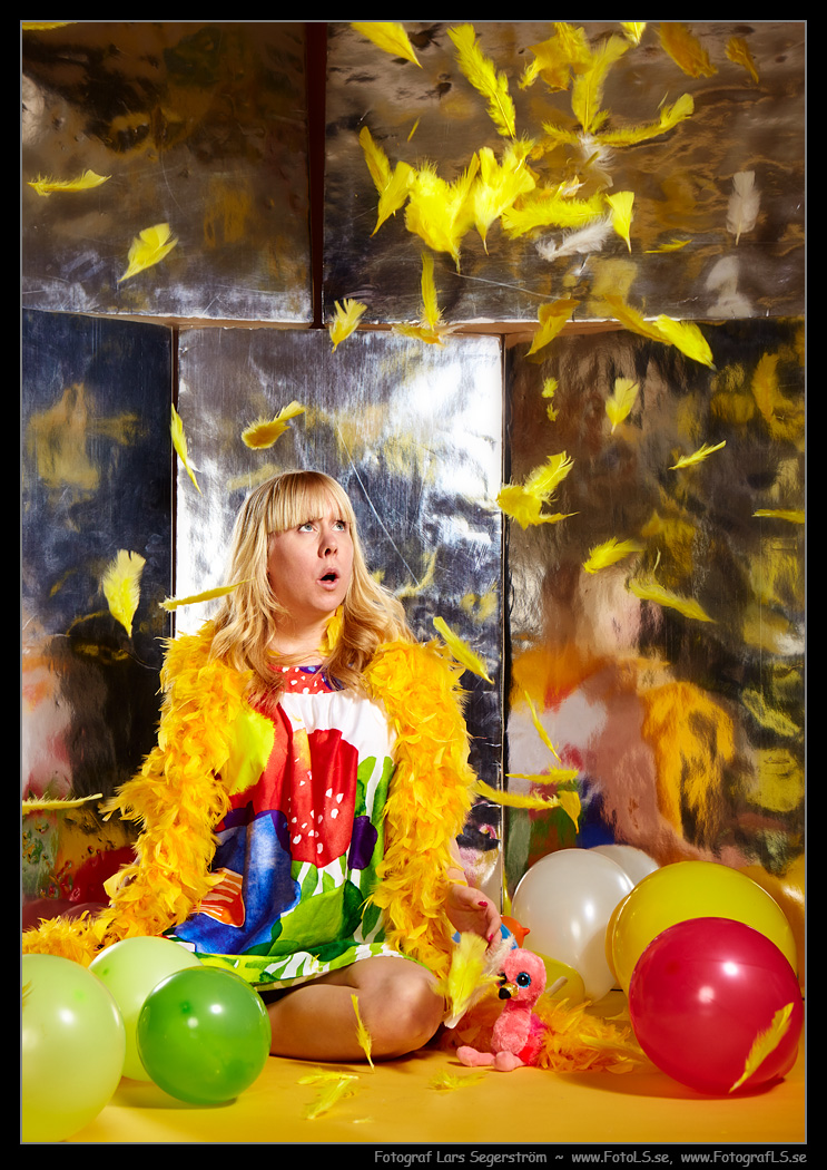 Färgglad bild av modell i gul fjäderboa bland ballonger och gula fjädrar.
