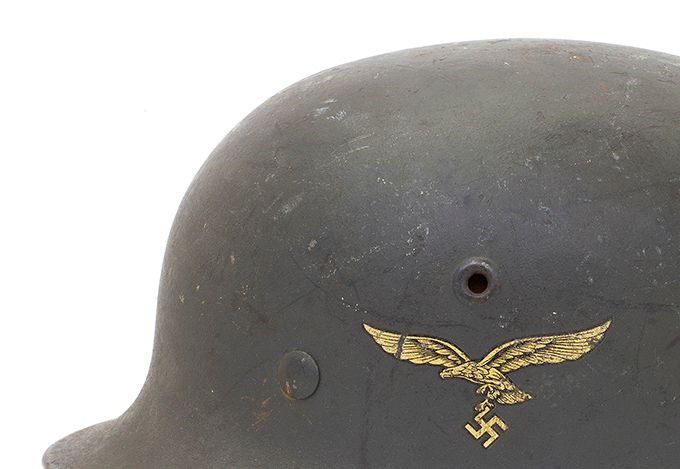 The M40 was produced from 1940 until mid-1942 (with the exception of the Quist factory) and as can be seen, the air vents are pressed into the sides of the shell during the manufacturing process. The helmet shown is a Q64 and has a an eagle spreading its wings in flight and clutching a swastika. This helmet is a Luftwaffe helmet (the German Air Force) and as was typical of M40 helmets, has insignia on the left side only. A Wehrmacht directive in March 1940 mandated that the national shield on the right side of the helmet be removed. From that point on, German helmets featured insignia on the left side only, with Police Troops being the exception. The smooth shiny paint was replaced by dull, textured paint. The crown is slightly more rounded in shape compared to the M35. The helmet shown has Luftwaffe mid-blue textured paint. From late 1940 the zinc-coated brass rivets were replaced by zinc-coated steel rivets which provided more strength.
 