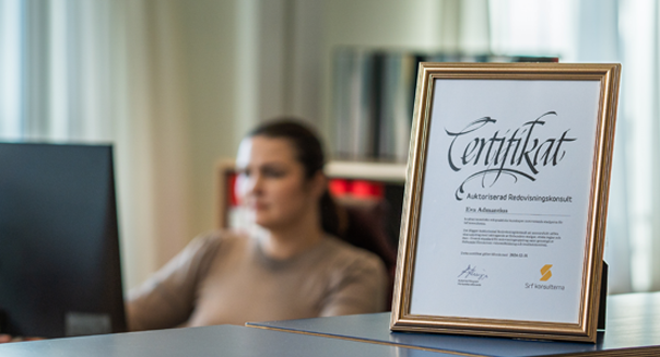 Bokföring - Redovisning - Ett inramat certifikat. Bakom certifikatet sitter en kvinna och jobbar vid datorn.