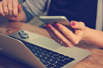 A person with pink nails holding a mobile phone in one hand with a laptop in the foregound